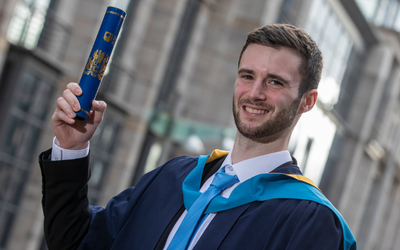 OU graduate Adam Hall pictured in a graduation gown and holding a degree scroll. 