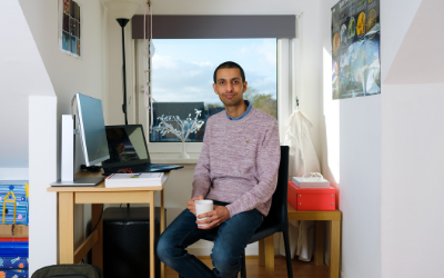 Open University graduate Ali seated at a table with a laptop, and holding a mug 