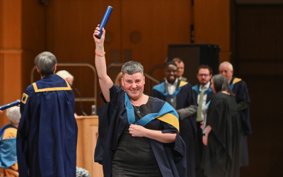 An OU graduate walking across the stage at the Glasgow Royal Concert Hall, holding up a graduation scroll. 