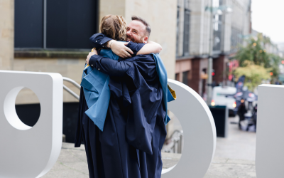 Two students in their graduation robes embracing in front of a large OU sign