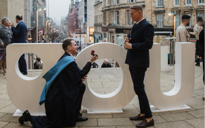 Steven Bell, wearing graduation robes and holding up a ring box to Clark Edgar, in front of giant letters OU.