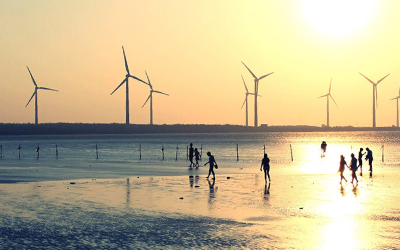 People walking on a beach in the foreground, with coastal wind turbines in the distance 