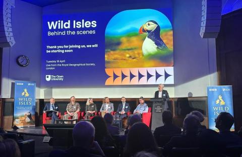 Stage showing five people seated and the Vice-Chancellor standing behind a podium. The stage is under a large screen with the title Wild Isles and an image of a puffin.