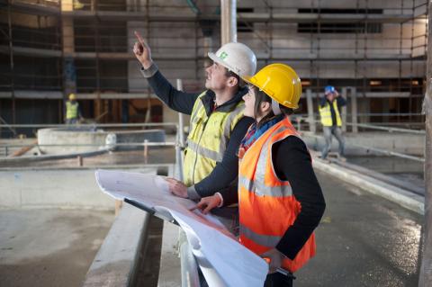 man and woman (in safety apparel) at construction site; man pointing ahead at something 